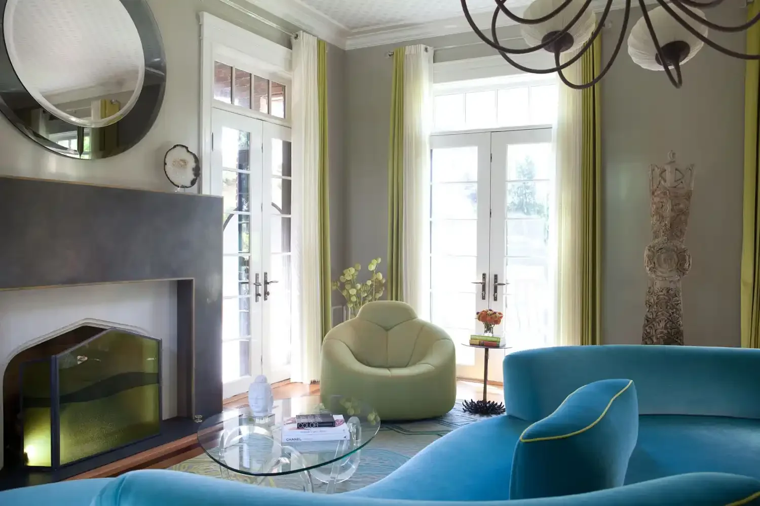 Living room with large windows, green curtains, a blue sofa, a cream chair, a glass coffee table with books, a fireplace, a mirror, and decorative items.