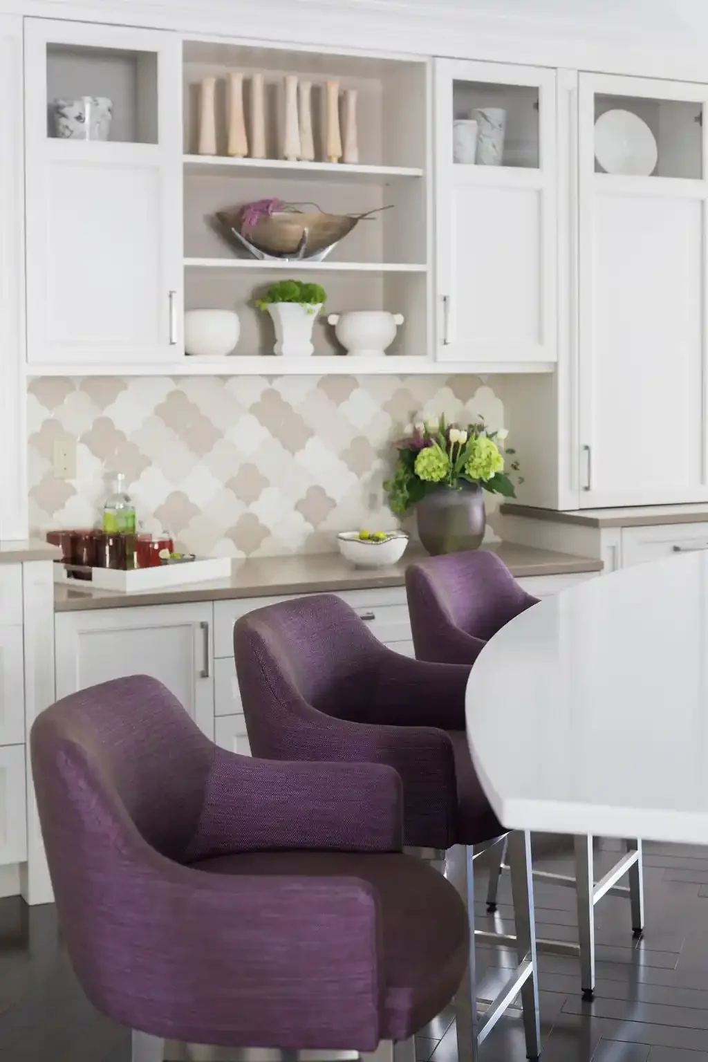 A modern kitchen with white cabinetry, purple chairs around a white table, decorative bowls, flowers in a gray vase, and a patterned backsplash.