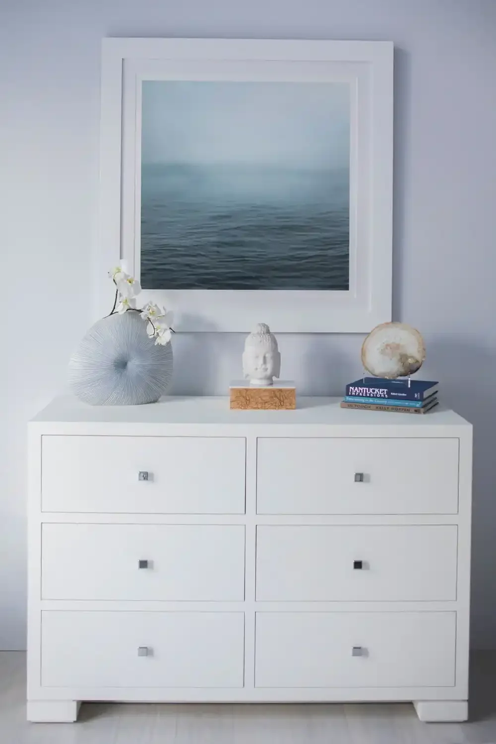 A white dresser with six drawers, decorated with vases, a sculpture, and books, with a large framed ocean photograph on the wall behind.