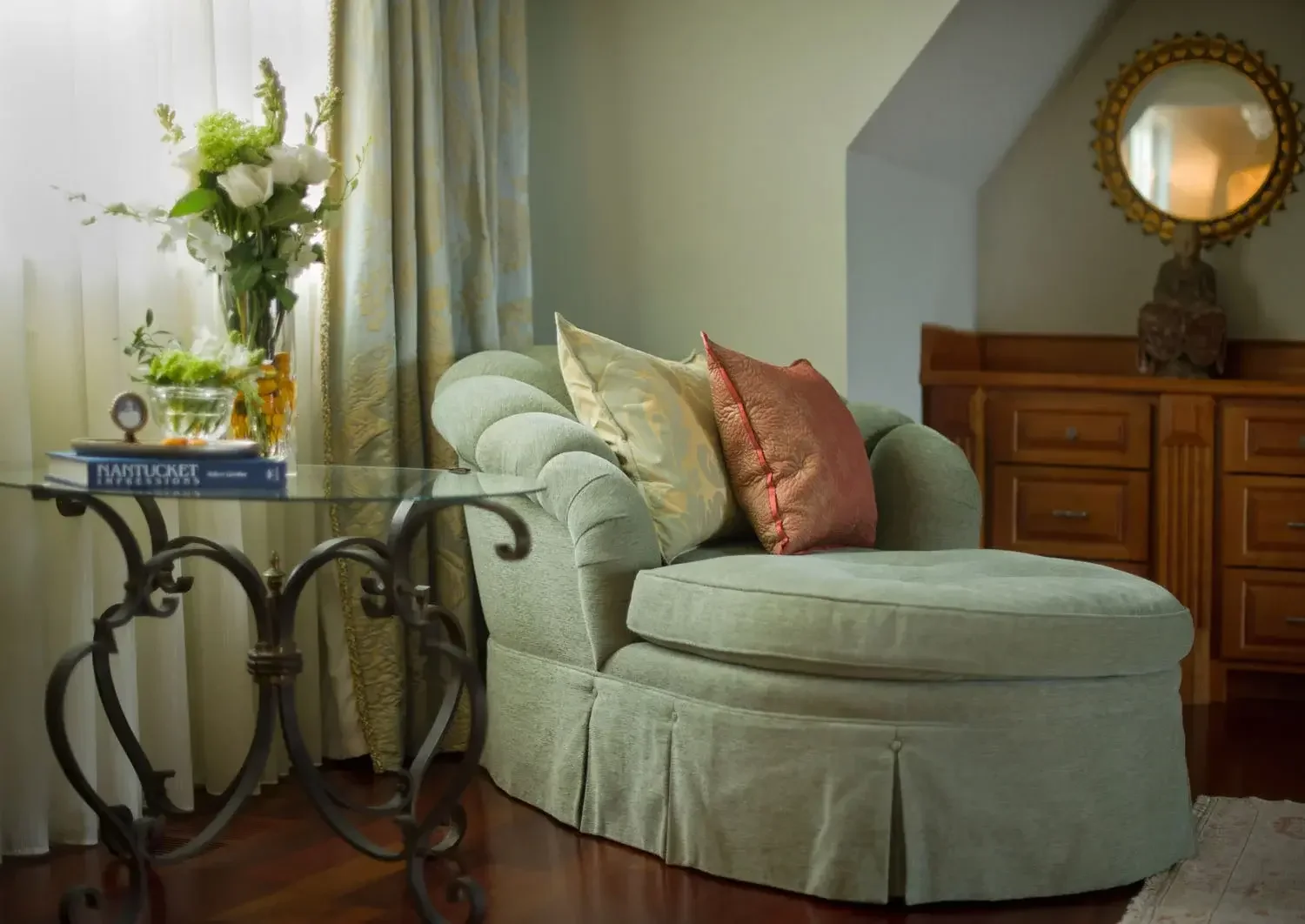 A cozy living room corner with a green upholstered chaise lounge, decorative pillows, a glass-top side table with a book, a vase of flowers, and a wooden sideboard with a round mirror above.