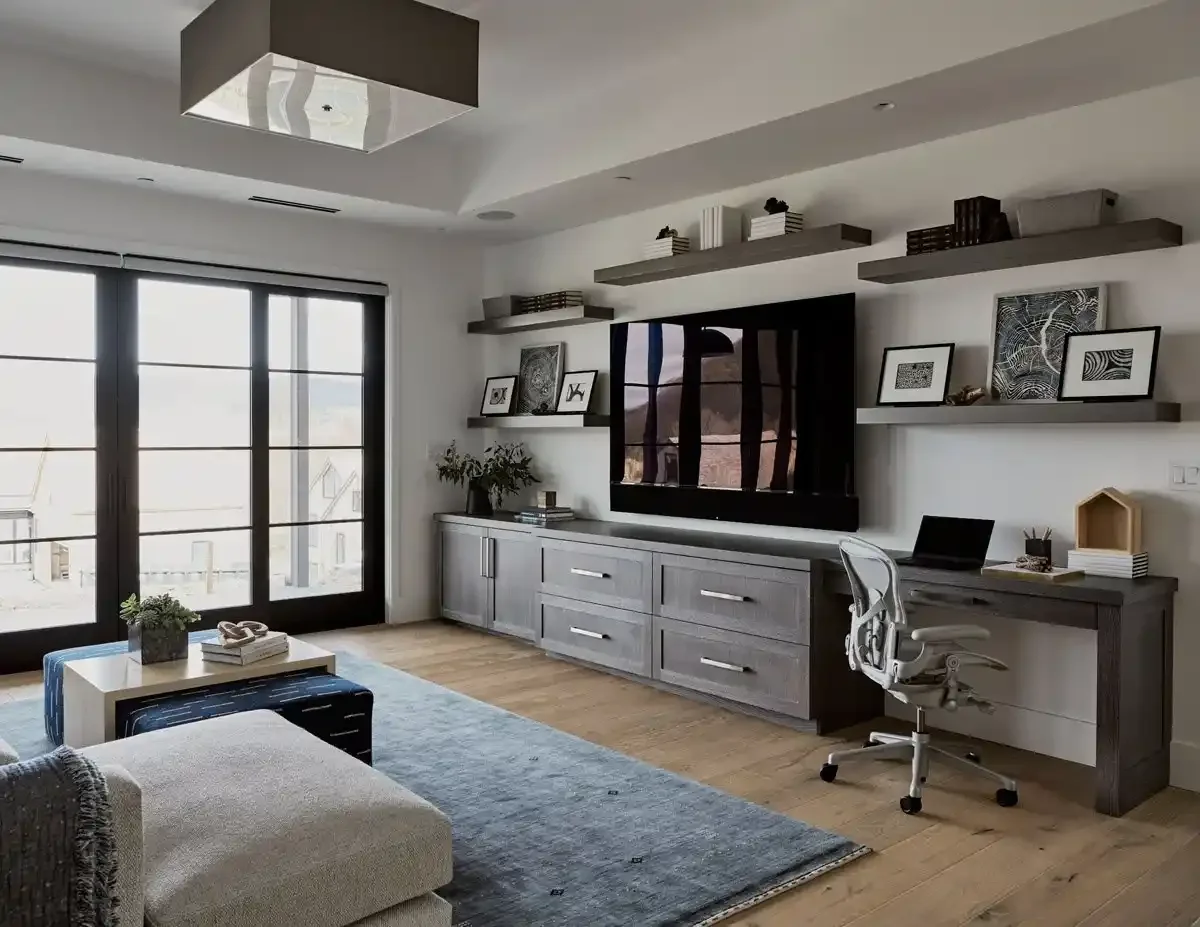 Modern living room with large glass doors, gray furniture, floating shelves with books and artwork, a mounted flat-screen TV, a desk with a white office chair, and decorative plants.