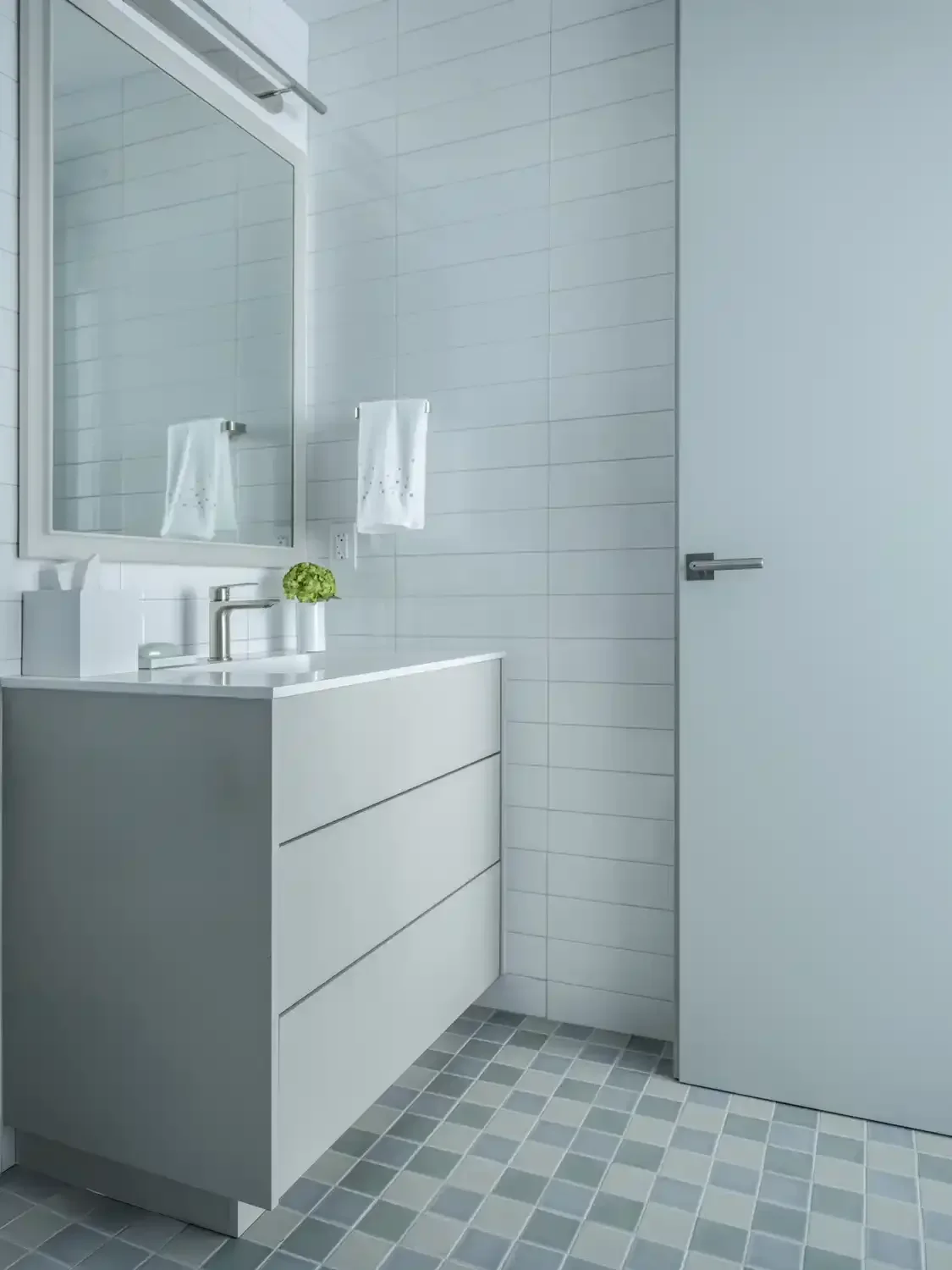Minimalist bathroom with white vanity, a large mirror, a small green plant, a soap dispenser, and a white hand towel on a rack. The walls are tiled in light gray, and the floor has small square gray tiles. There is a white door with a modern handle.