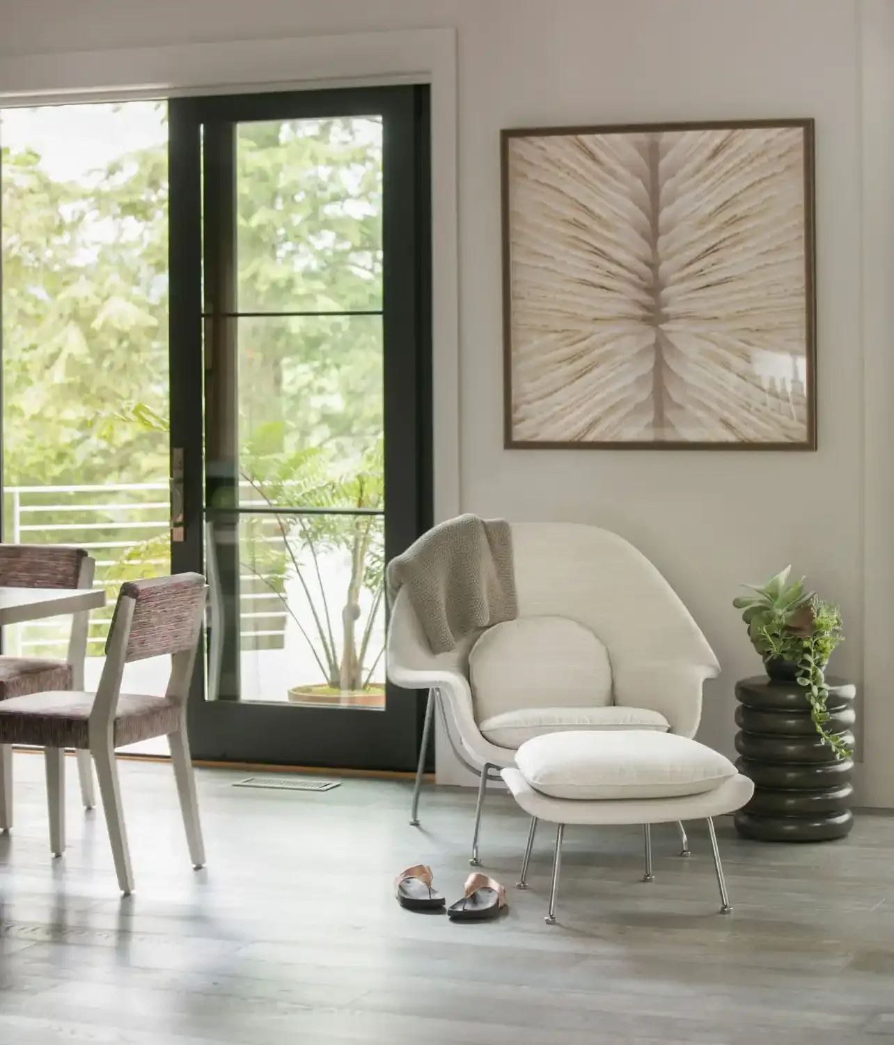 Living room with white armchair and ottoman, potted plant, large artwork on wall, glass door leading outside, and dining area with chairs.