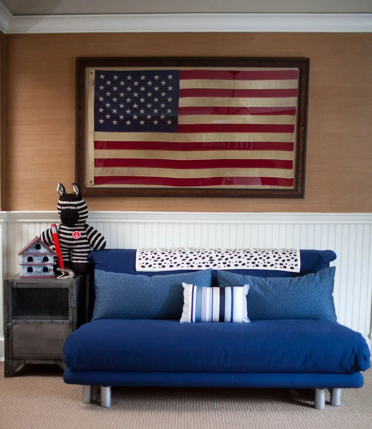 A living room with a framed American flag on the wall, a blue sofa with a striped pillow, a small stuffed zebra with a peace sign on its back, a red and gray birdhouse, and a small side table.