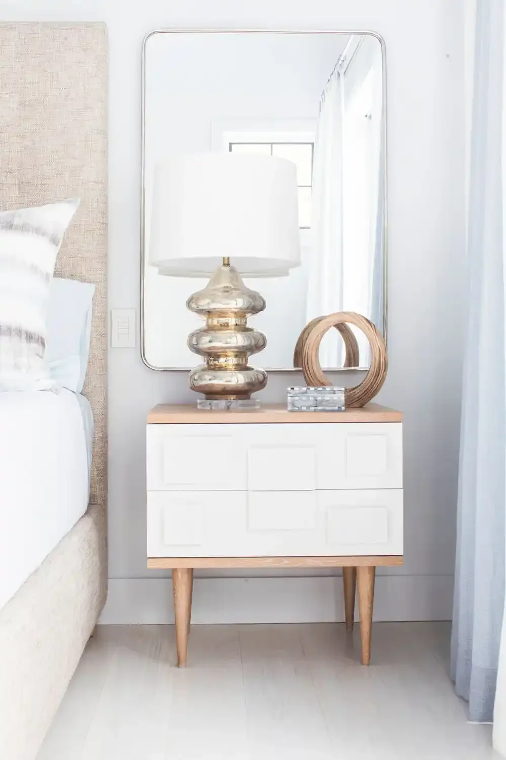 Decorative bedroom nightstand with a modern glass table lamp, wooden sculpture, and a mirror behind.
