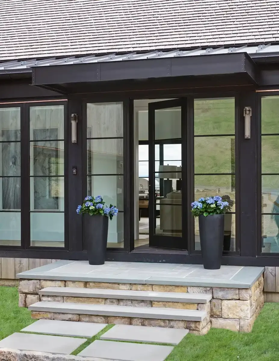 Front entrance of a house with a black-framed glass door and large windows, two tall black planters with blue hydrangeas, stone steps, and a grassy yard outside.