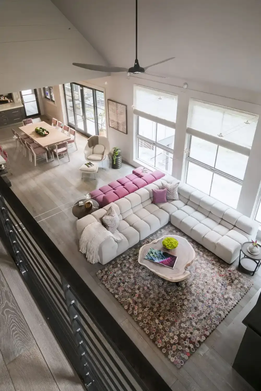 Overhead view of a bright, spacious living room with large windows, a white sectional sofa with purple accent pillows, a coffee table with a decorative bowl and magazine, and a multicolored rug. Adjacent dining area with a wooden table and pink uphol