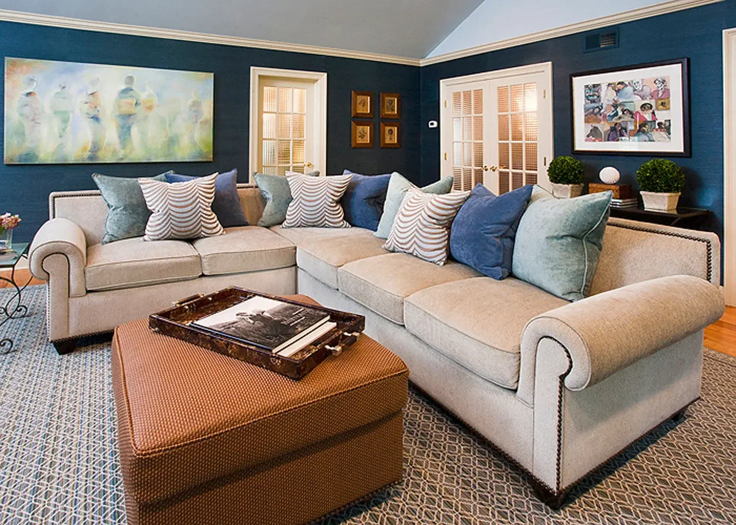 Living room with beige sectional sofa and patterned throw pillows, dark blue walls, framed artwork, window with blinds, coffee table with books, patterned rug, and decorative plants.
