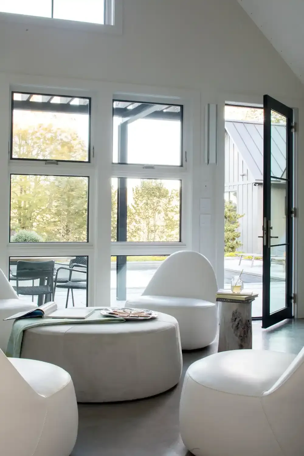 Bright, modern living room with large windows, white furniture, and an open door leading outside to a patio with trees and outdoor furniture.