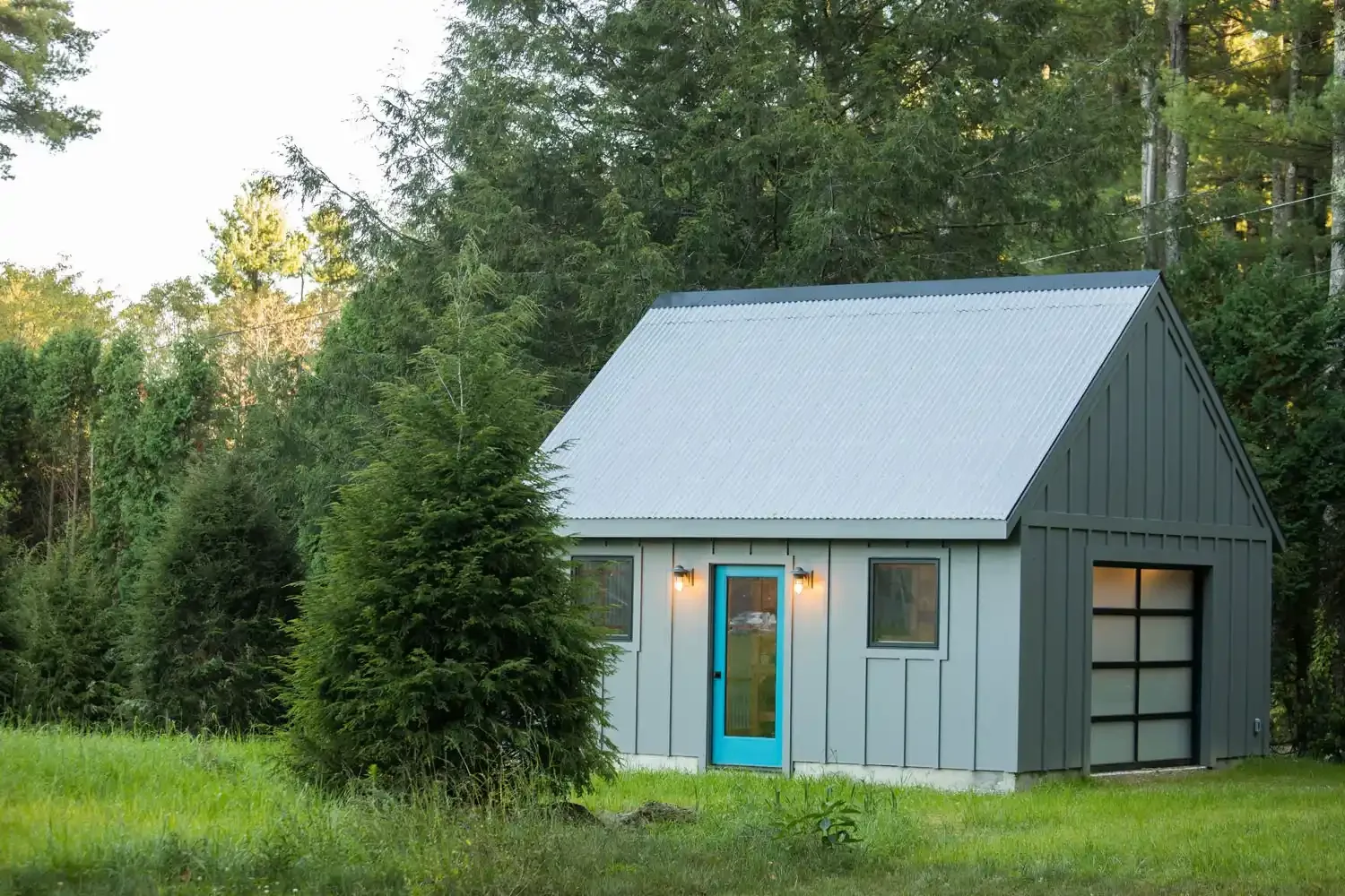 Small gray shed with a blue door, two exterior lights, a garage door, surrounded by greenery and trees.