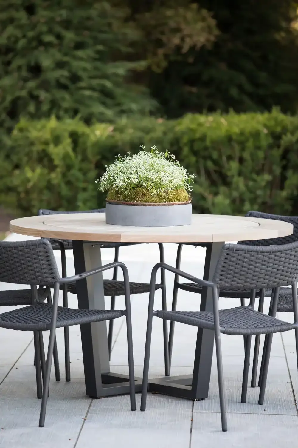 Outdoor patio table with four black woven chairs surrounding it, and a large metal planter with white flowers as centerpiece, against a background of green bushes and trees.