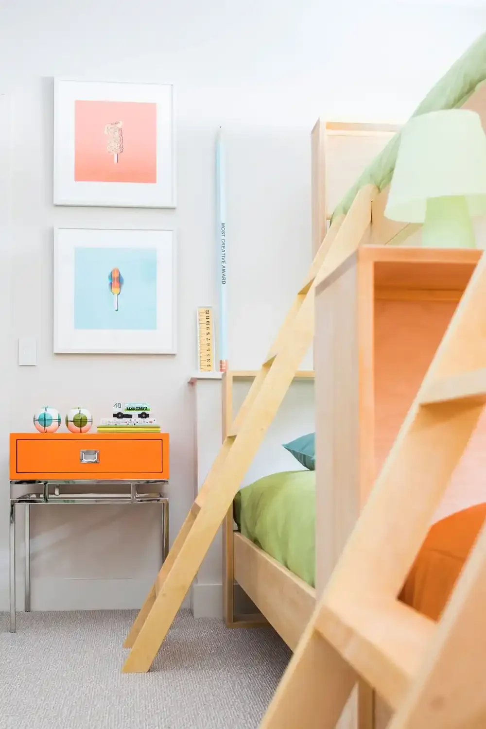 Interior of a children's bedroom with a wooden bunk bed, green bedding, and a colorful side table with toys and books, decorated with modern art prints on the wall.
