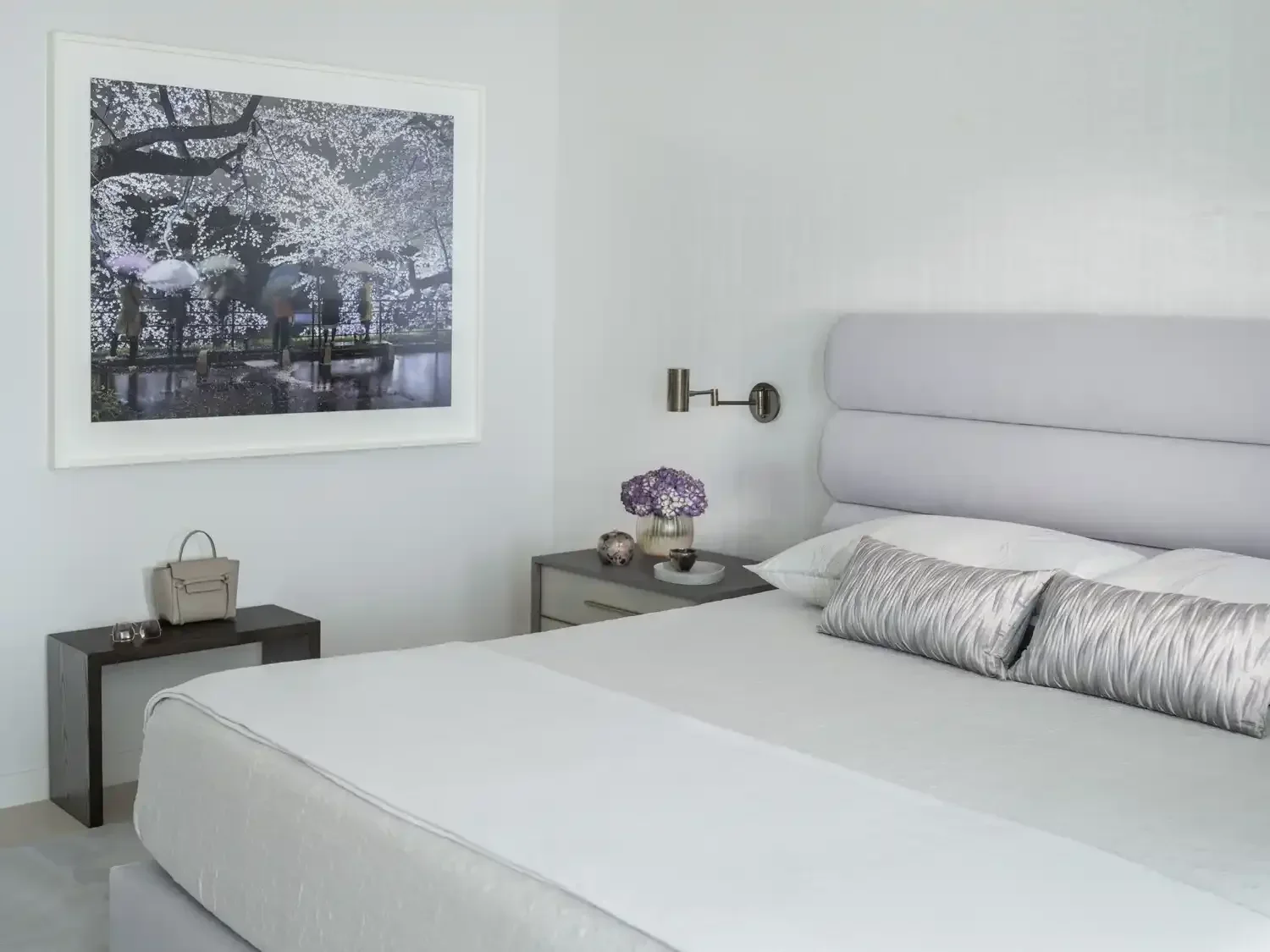 A modern bedroom with a white bed, silver pillows, and a gray bedside table with a vase of purple flowers, alongside a framed picture of cherry blossoms on the wall.