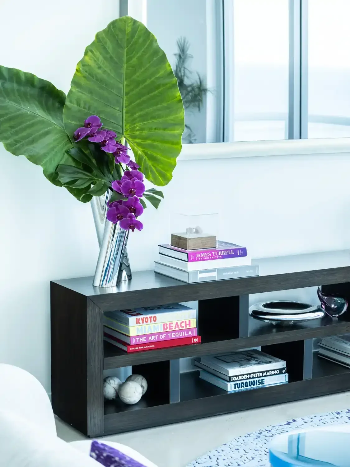 Decorative black shelf with books, a silver vase with large green leaves and purple orchids, and decorative stones in a modern, bright room.