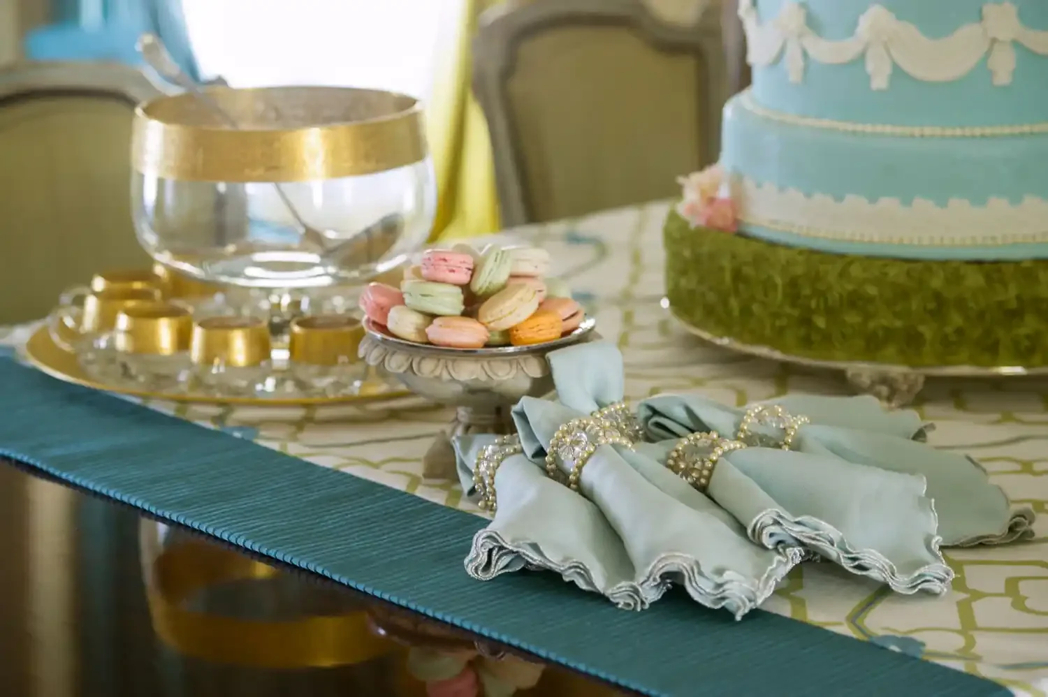 A decorated table with pastel-colored macarons on a silver dish, a large blue cake with white decorations on a stand, a glass bowl with a gold rim, and a pile of pearl jewelry and light blue napkins with pearl rings, set for a celebration or dessert 