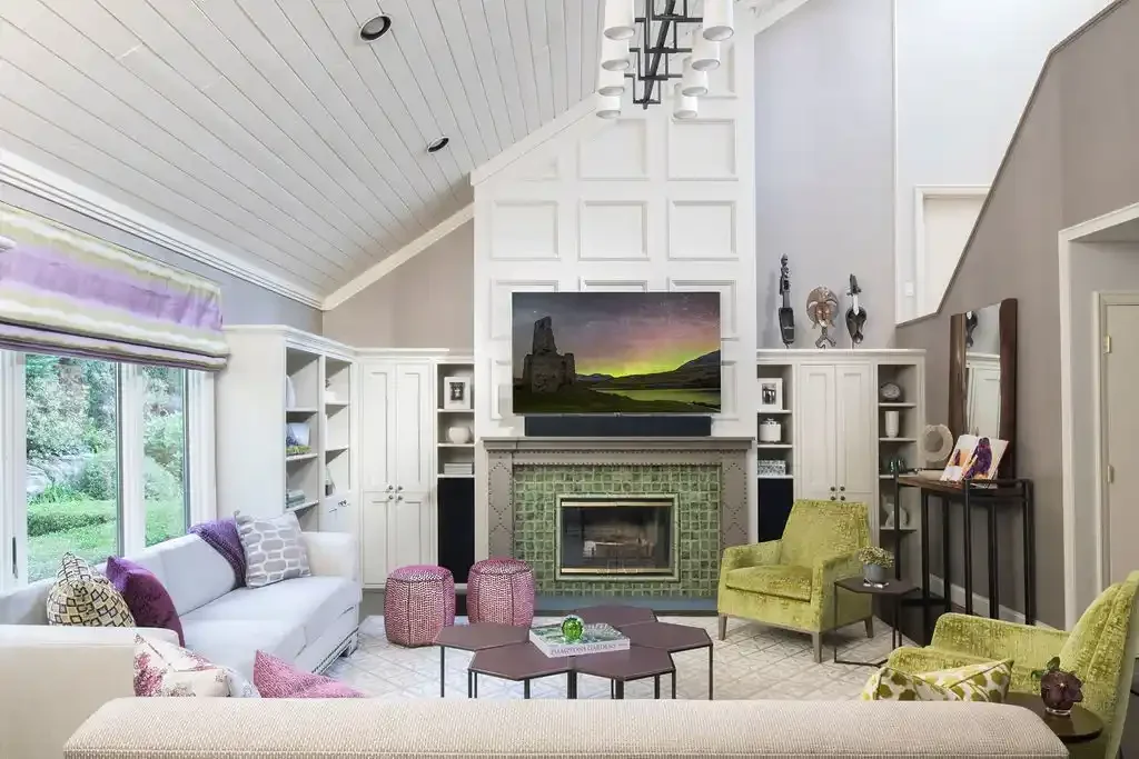 Living room with vaulted ceiling, white walls, a fireplace with green tiles, a large flat-screen TV above, and assorted seating including a white sofa and green armchairs. There's a large window with purple accents on the left.