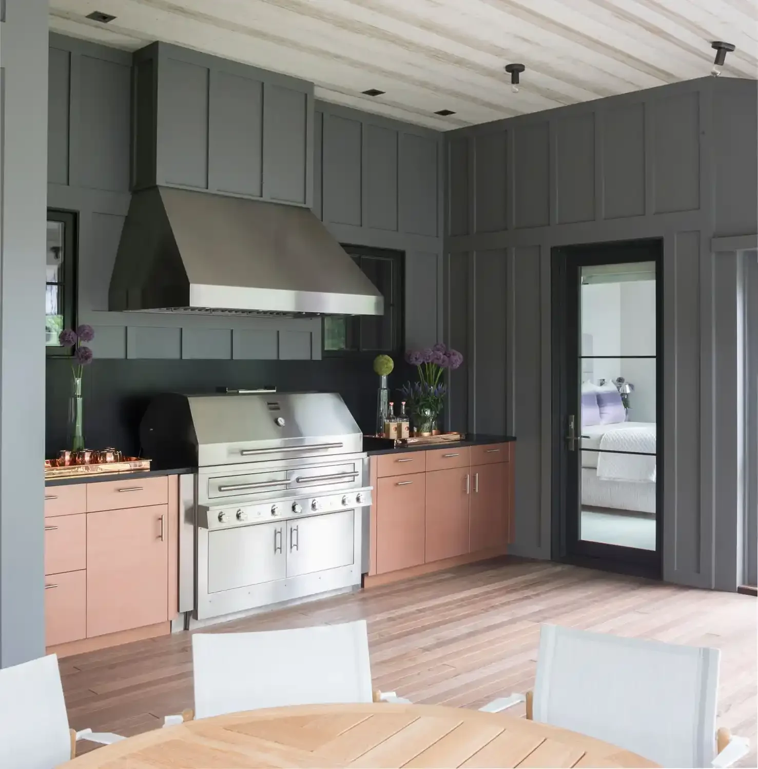 Modern kitchen with gray cabinets, a stainless steel range hood, a silver grill, pink lower cabinets, and decorative flowers on the counter. There's a black door with glass leading to a bedroom.