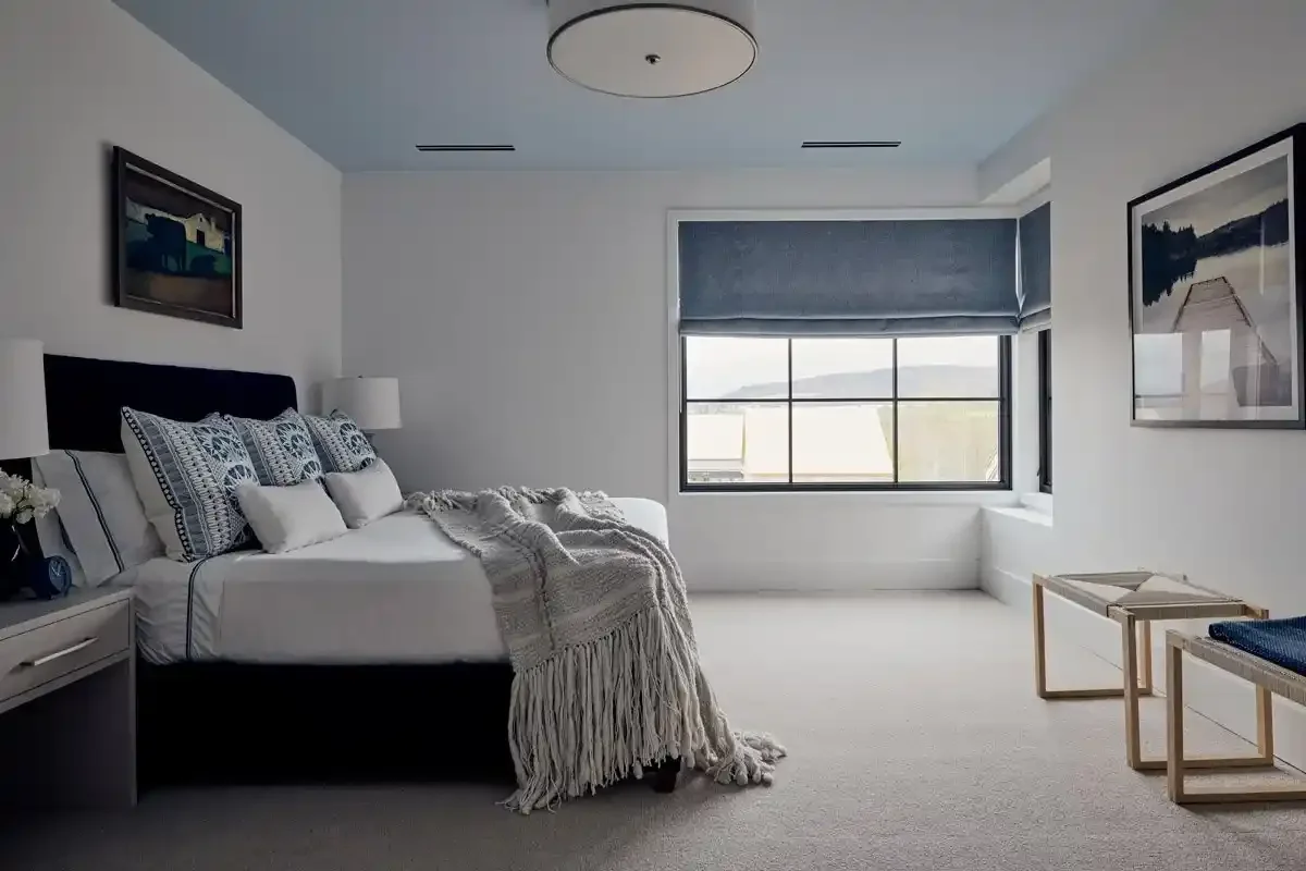  Bedroom with a large bed, white bedding, patterned pillows, throw blanket, nightstand with lamp and flowers, large window with a blue valance, framed artwork on white walls, and a small bench at the foot of the bed.
