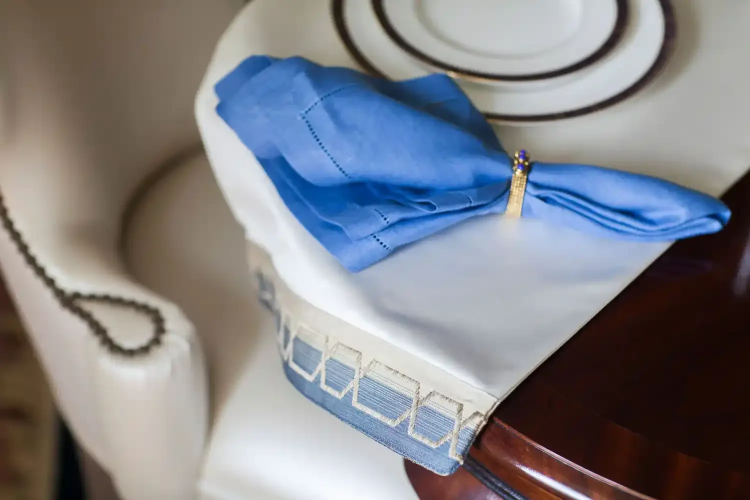 A blue fabric bag with a gold band, resting on a white tablecloth with a decorative border, next to a cream-colored telephone with a cord, on a wooden table.