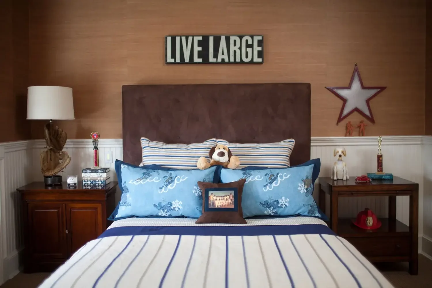 A neatly made bed with a brown headboard, decorated with stuffed animals and pillows, flanked by two wooden nightstands with lamps and decorative items, a wall sign reading 'LIVE LARGE' above the headboard, and patriotic decorations including a star,