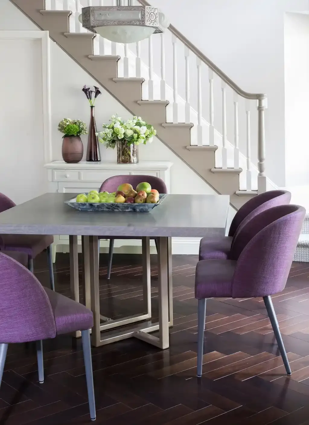A dining room with a gray table, surrounded by purple chairs, on a dark hardwood floor. There is a white sideboard with vases and flowers underneath a staircase with white balusters, and a bowl of apples on the table.