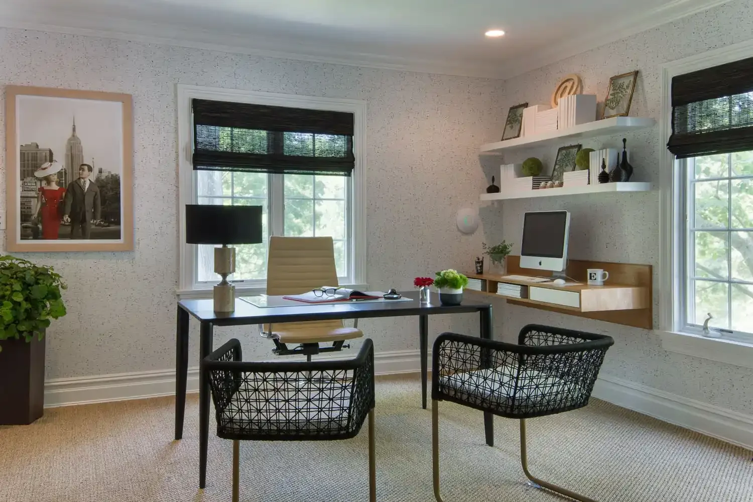 Home office with beige carpet, black desk, beige chair, black wicker chairs, window with black blind, white walls with textured pattern, white shelves with books and decor, computer, plants, framed artwork, and table lamp.