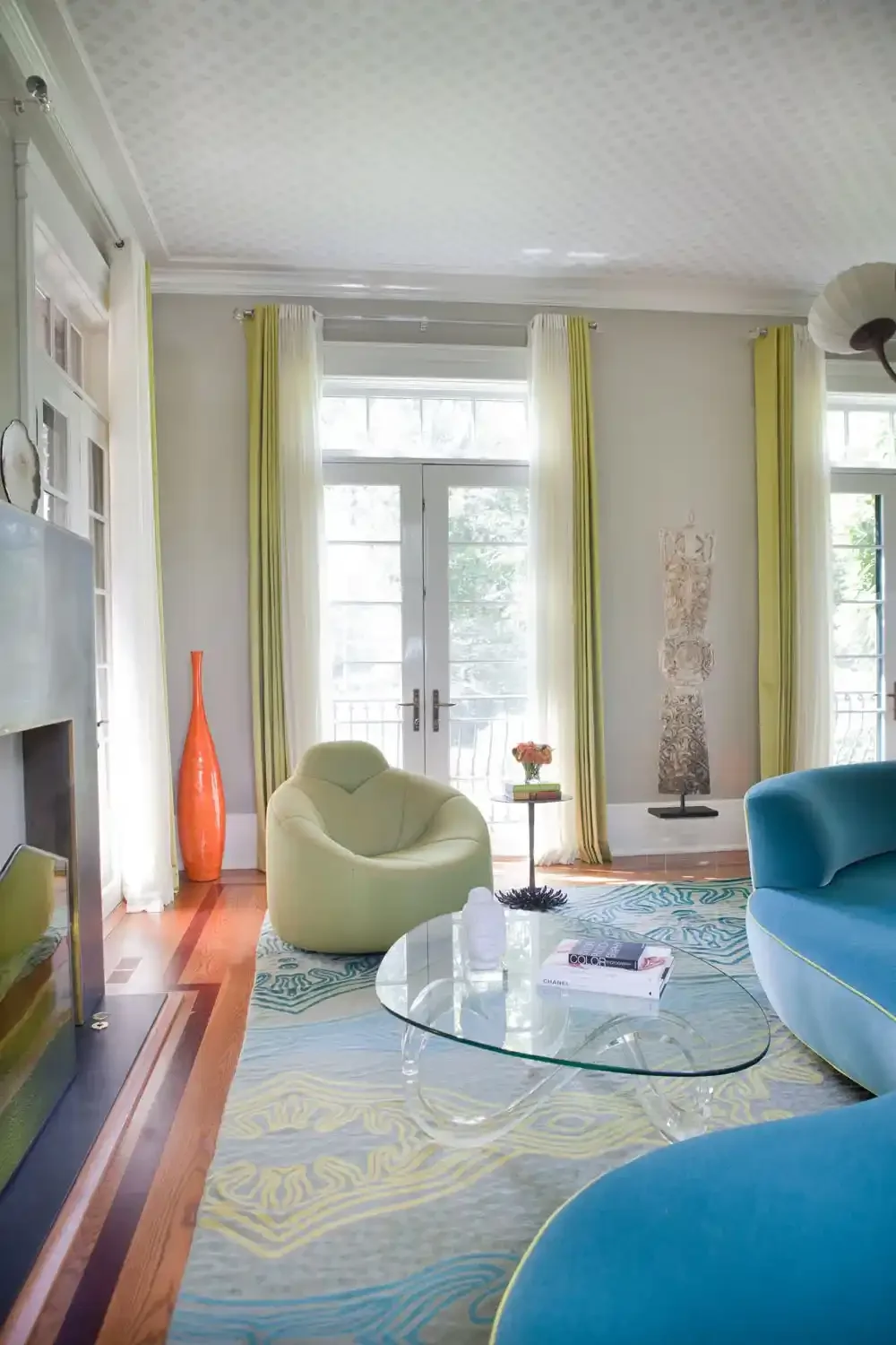 Bright living room with bay windows, yellow curtains, a glass coffee table, a green armchair, a blue couch, and decorative items, including an orange vase and wall art.