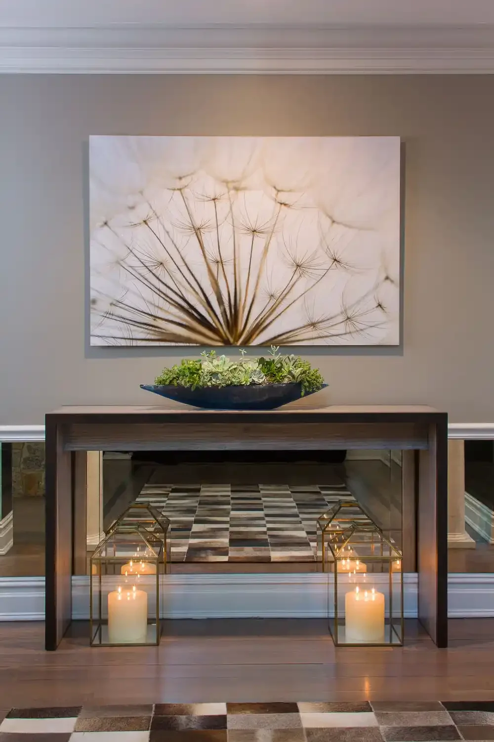 A framed artwork of a dandelion seed head hangs above a wooden console table with a black bowl filled with green plants. Two glass lanterns with lit candles are placed on the floor, flanking the table. The wall is painted beige, with a mirror on the 