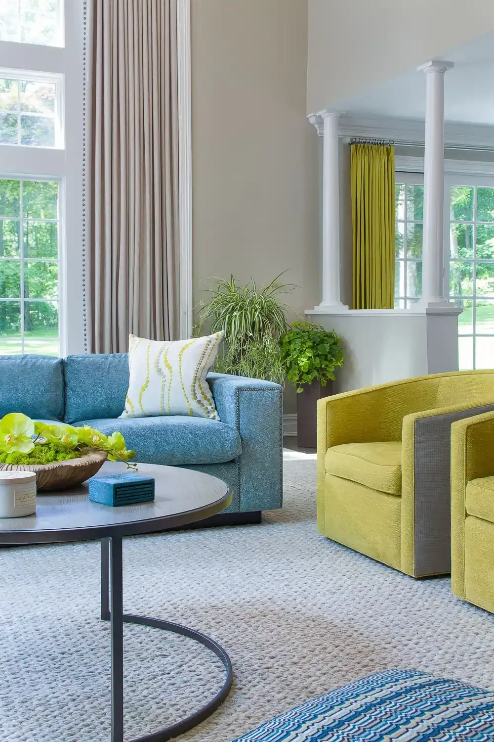 Bright living room with large windows, beige curtains, a light blue sofa with a patterned throw pillow, yellow armchairs, a round coffee table with decor, and green plants.