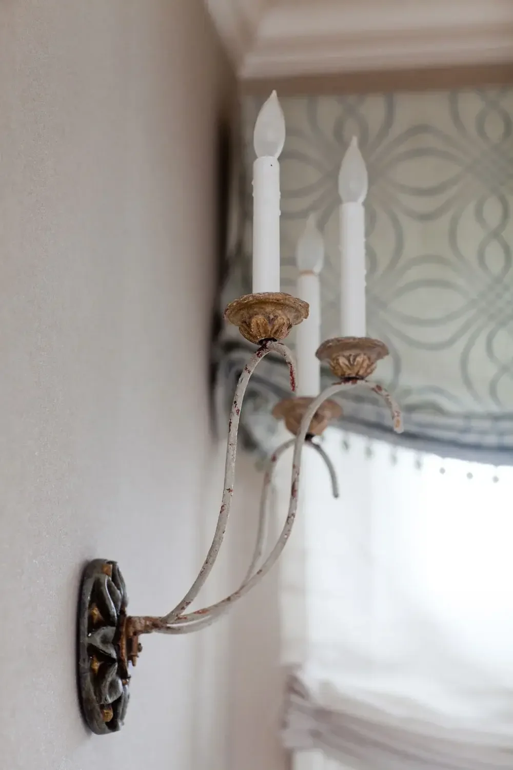 A vintage white wall sconce with three candle-shaped light bulbs, featuring a metal base that is rusted and worn, mounted near a window with a patterned shade.