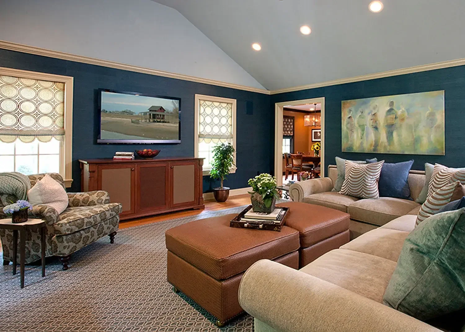 Living room with a blue accent wall, beige sofas with pillows, a patterned armchair, a wooden TV stand with a television, plants, and artwork on the walls. Adjacent dining room visible through an opening.