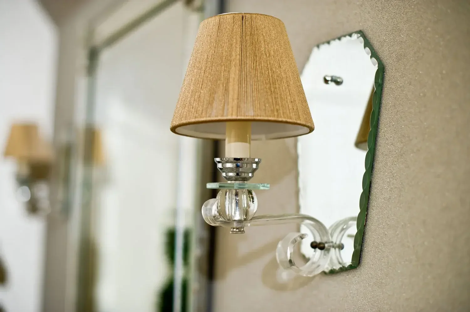 A wall-mounted bedside lamp with a beige lampshade, glass and metal design, reflected in a mirror on a textured wall.