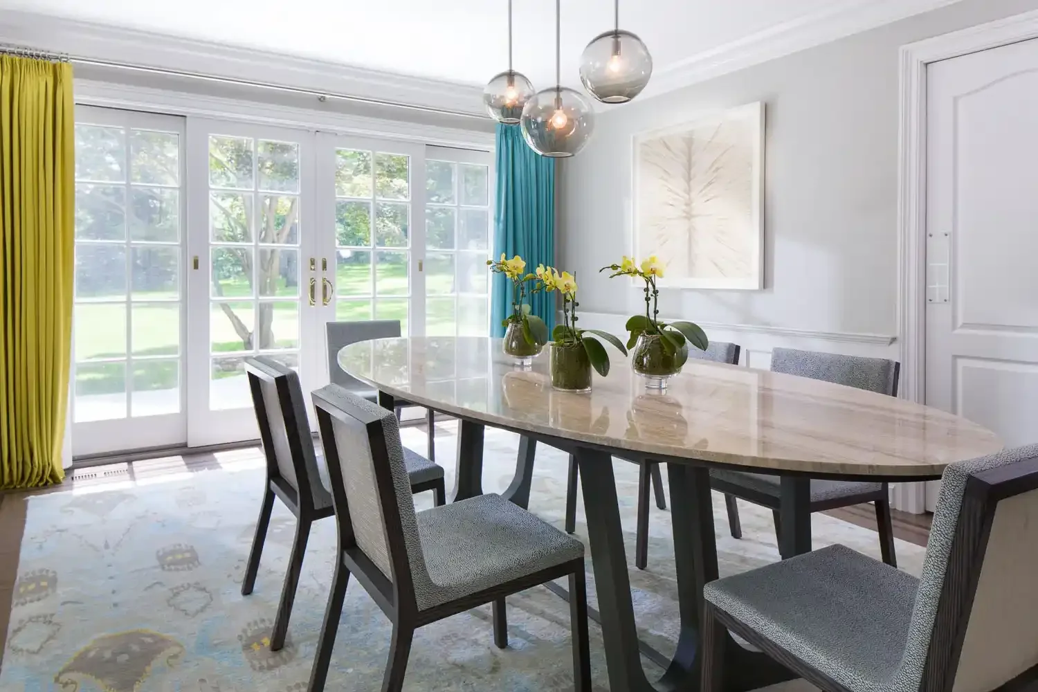 Elegant dining room with a marble-topped oval table, six upholstered chairs, yellow and blue curtains, a modern chandelier, and large windows showing a green outdoor space.