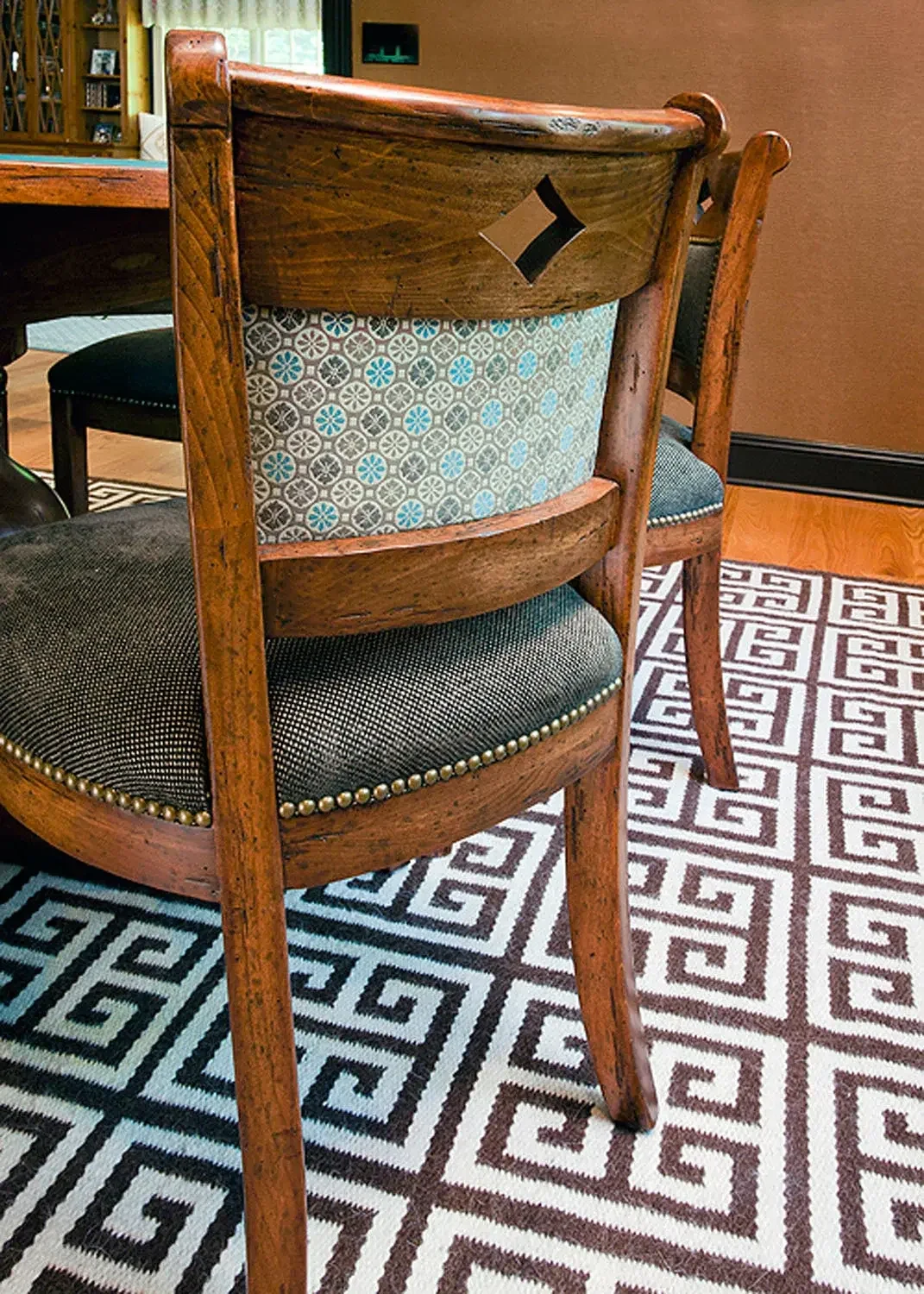 A wooden dining chair with a patterned upholstered backrest and a padded seat with nailhead trim. The chair is placed on a geometric patterned rug in a dining room.