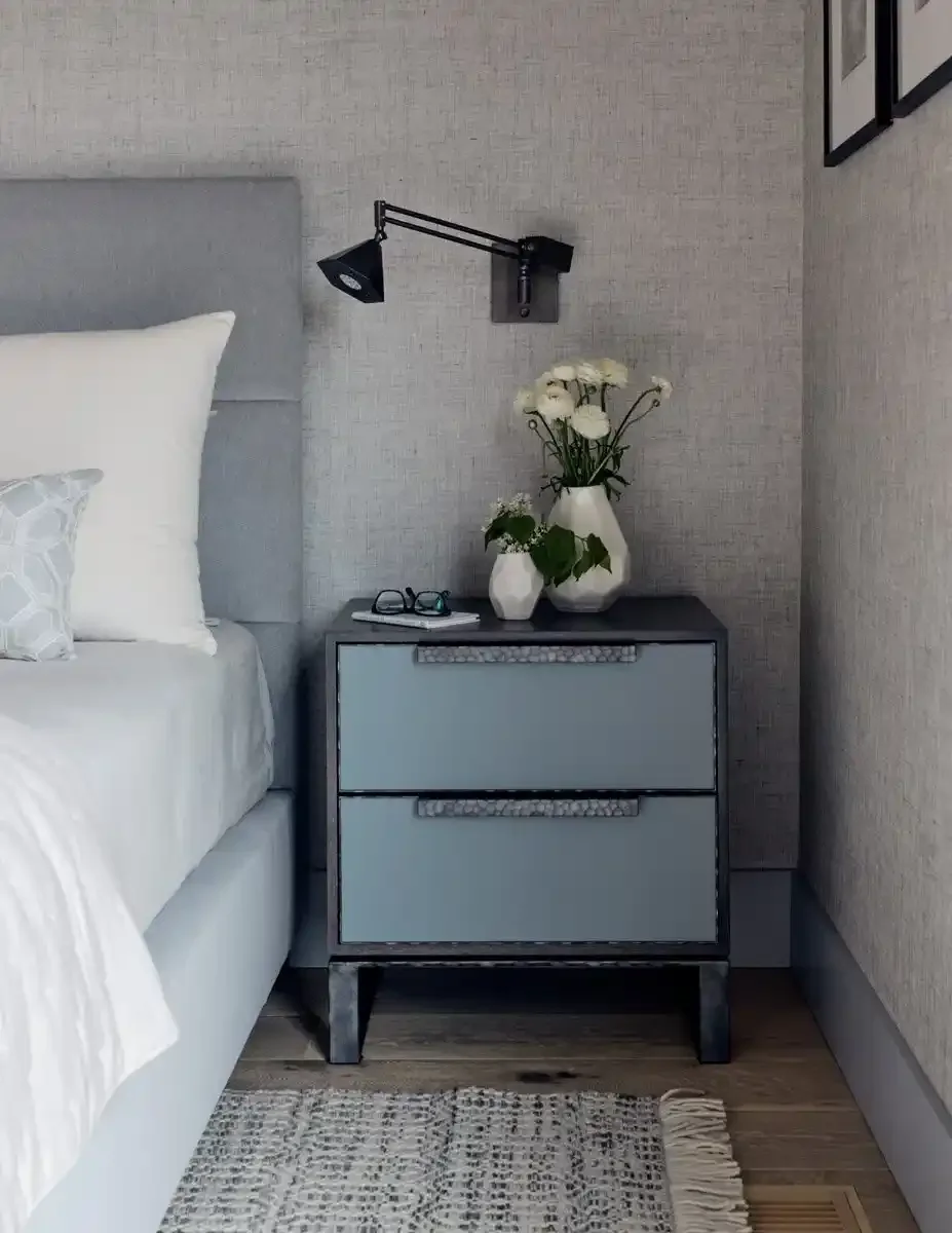 A nightstand with two drawers, a pair of eyeglasses, a book, and three white vases with flowers on top, next to a bed with white bedding, in a bedroom with textured gray walls.