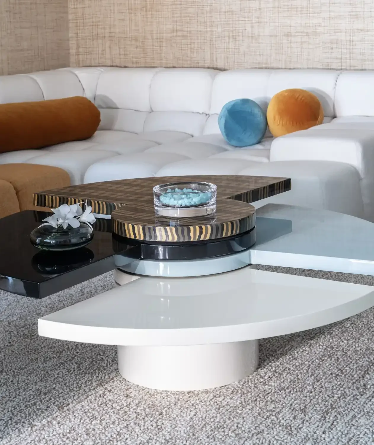 Modern living room with a white sectional sofa, decorative pillows, and a unique layered coffee table with wooden, black, and white surfaces, decorated with a small vase, flowers, and a glass bowl of blue decorative beads.