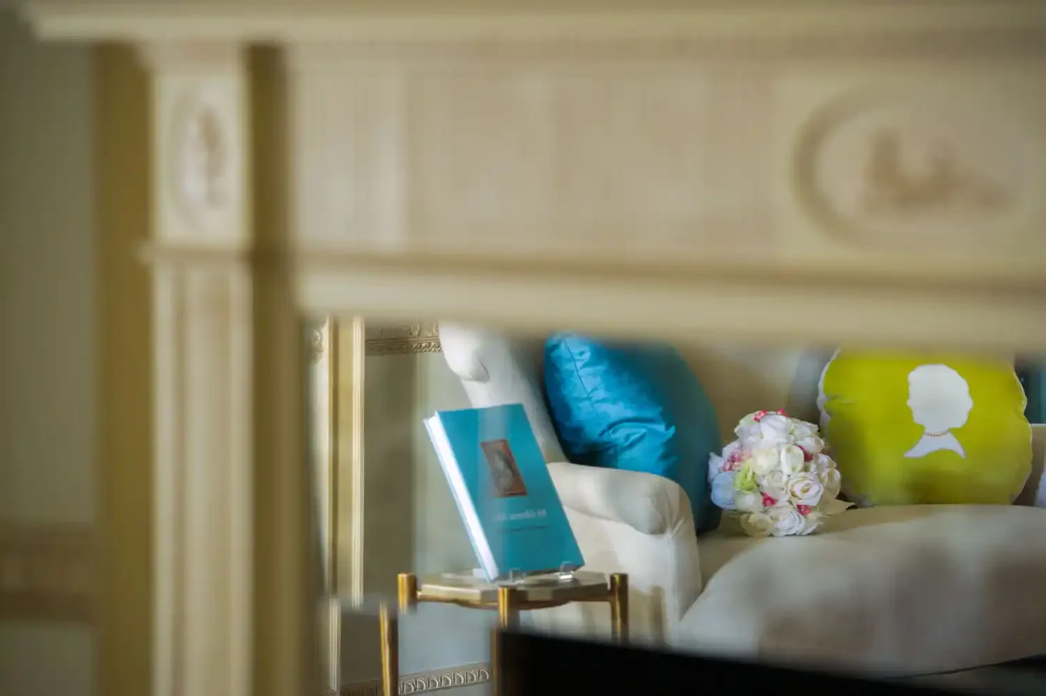 A view through a gap in furniture showing a sofa with blue and purple pillows, a bouquet of white and pink flowers, a framed picture on a stand, and decorative wall elements.