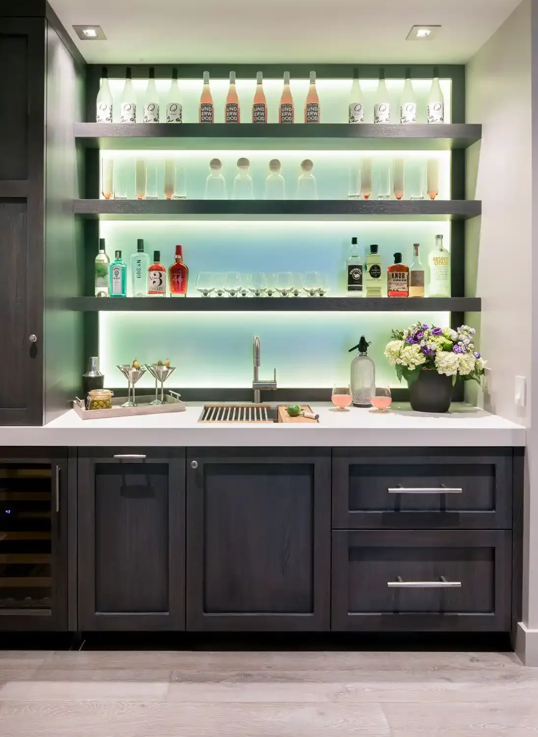 Modern home bar with dark wood cabinets, illuminated shelves with various bottles of alcohol, cocktail glasses, and a flower arrangement.