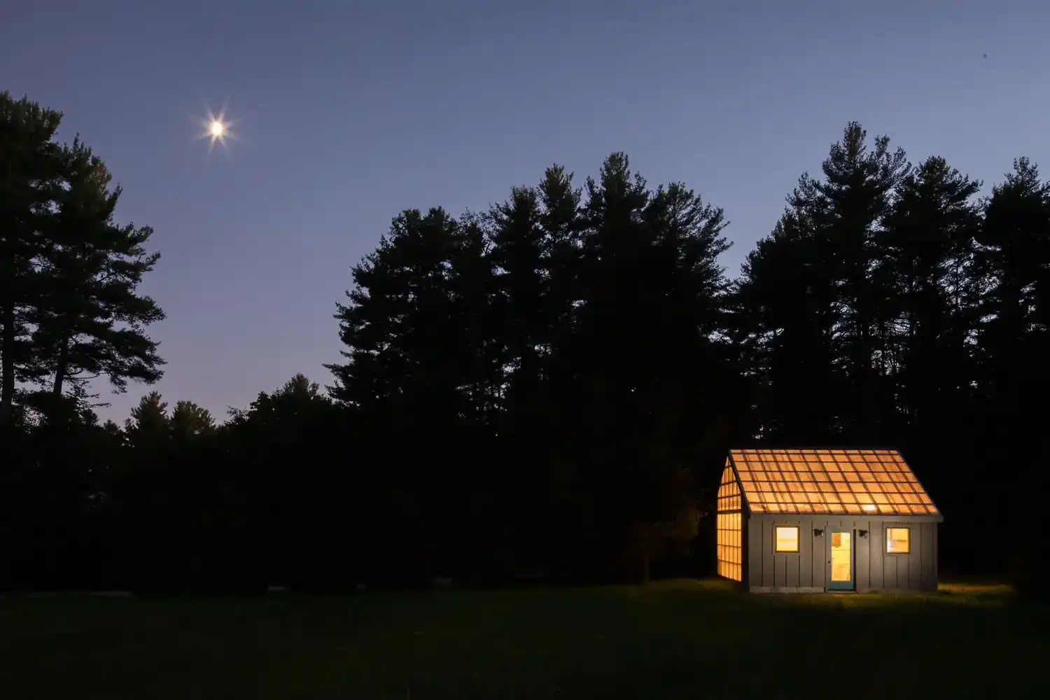 A small shed with a glowing interior at night, surrounded by dark trees and a clear sky with the moon visible.