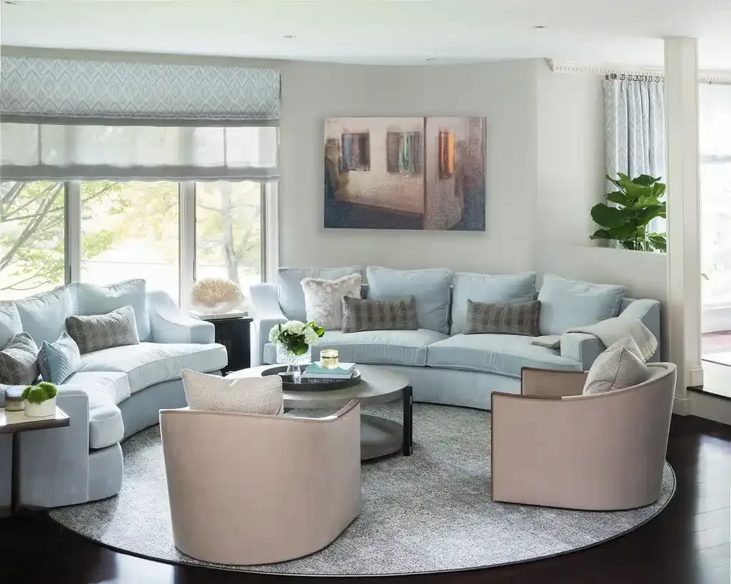 Bright living room with light blue sectional sofa, armchairs, a round coffee table, and large windows with white curtains and shades.