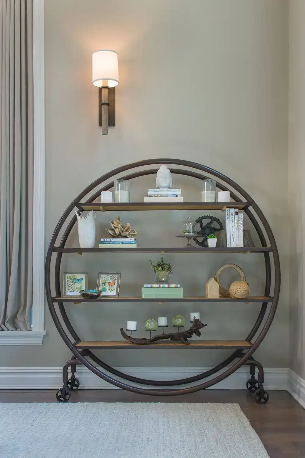 Decorative circular bookshelf with various books, plants, and decorative objects, against a beige wall with a wall sconce light above.