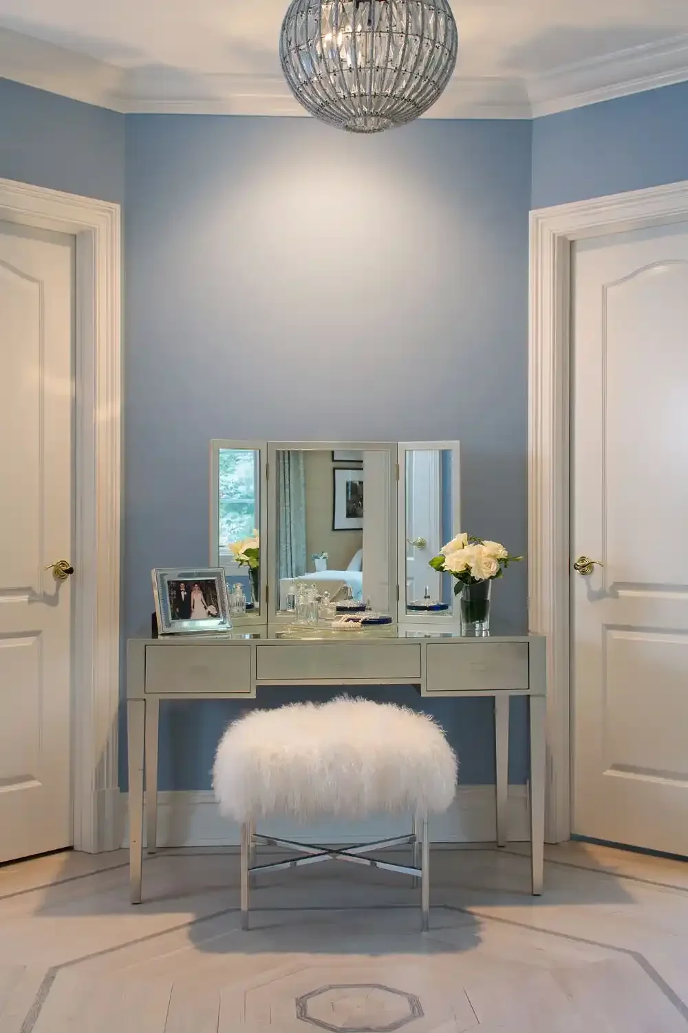 A bedroom with light blue walls, white doors, a silver vanity table with a mirror, a white fluffy stool, a framed photo, and a vase with white roses.