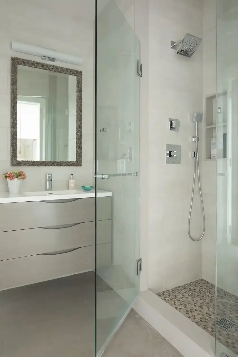 Modern bathroom with a glass shower enclosure, showerhead, handheld shower, and a vanity with a mirror and decorative flowers.