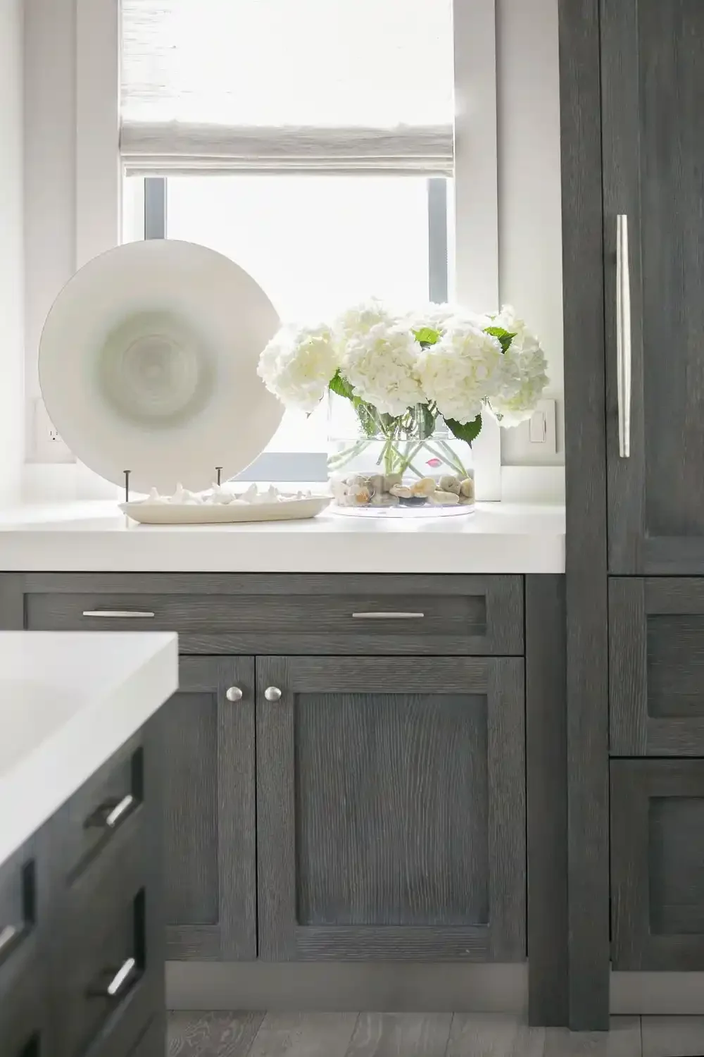 Kitchen window ledge with a large white plate, a white ceramic dish filled with white decorative objects, and a vase of white hydrangea flowers.