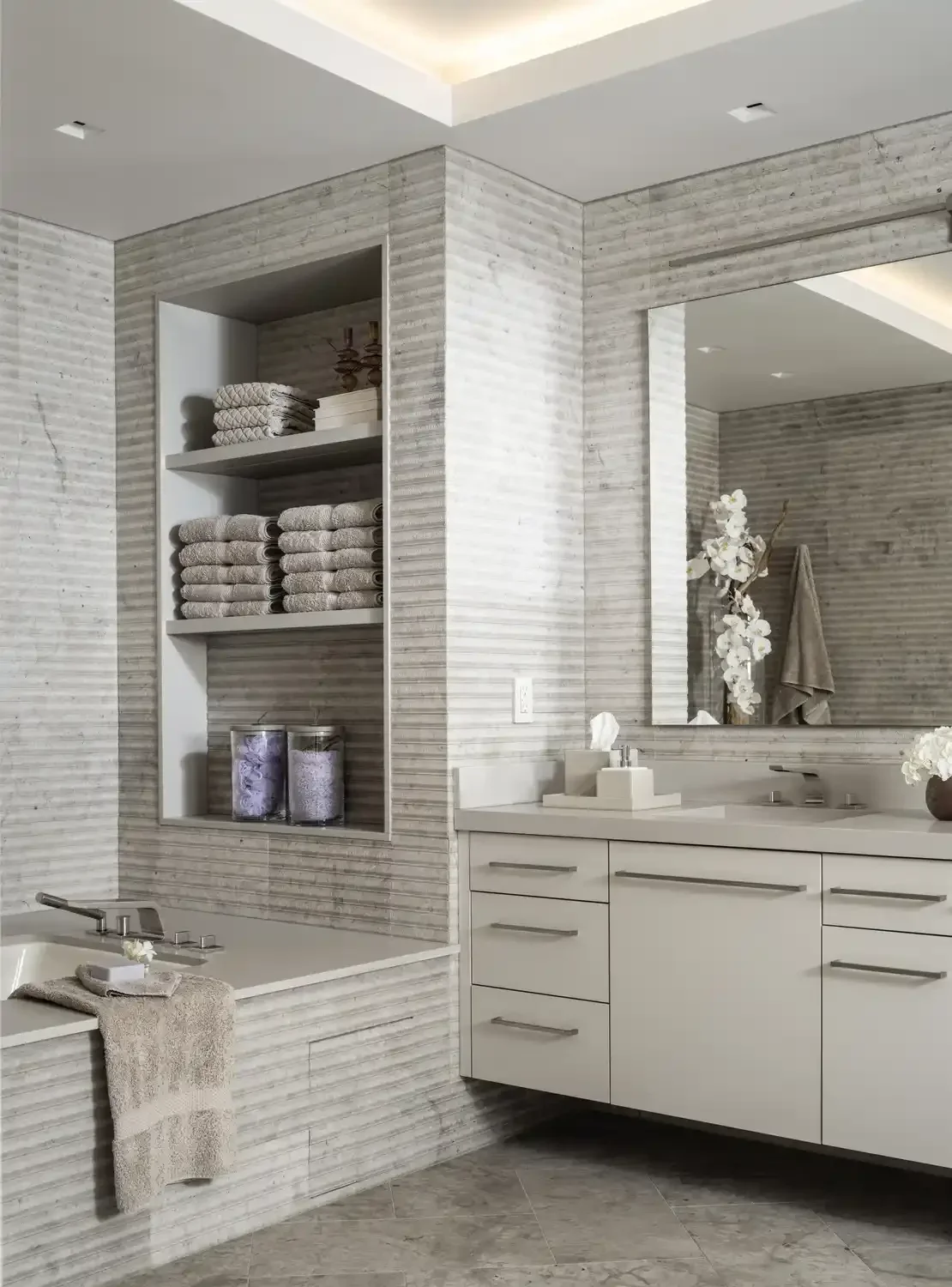 Modern bathroom with a built-in shelf holding folded towels and decorative jars, a white vanity with a large mirror, a sink, and a vase with white orchids.