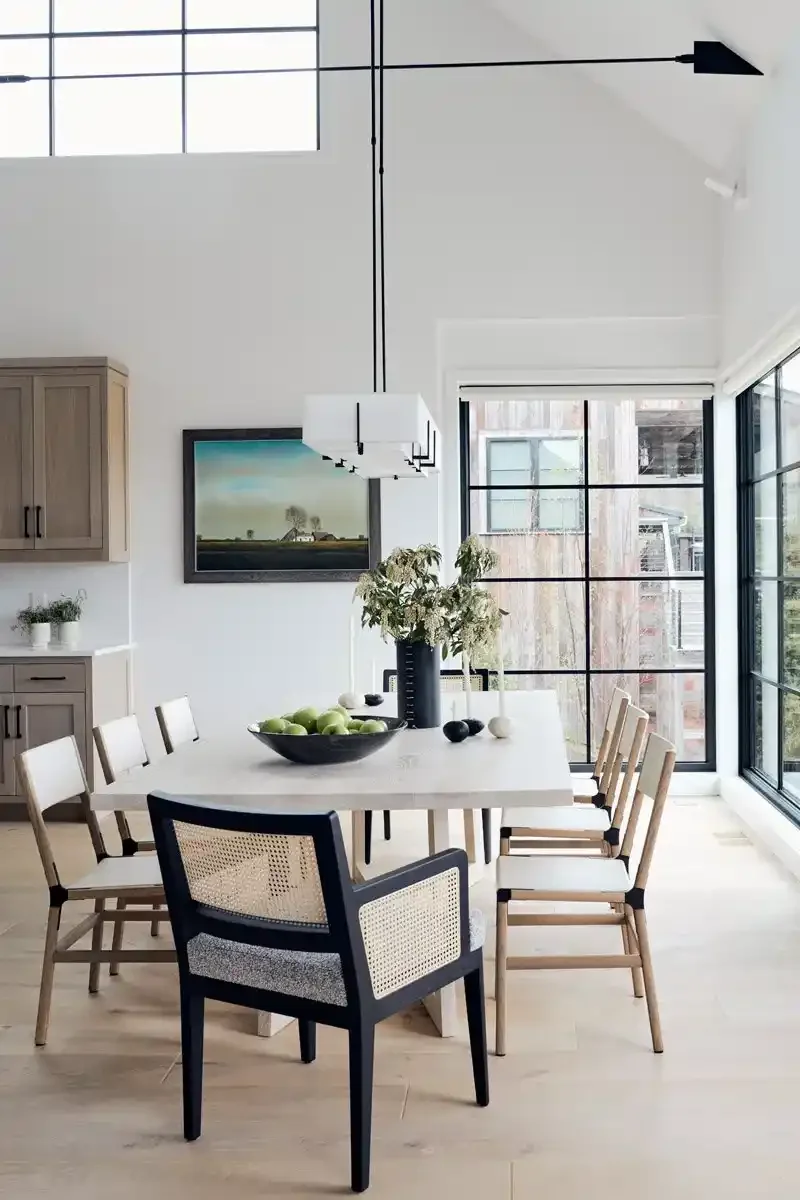 Modern dining room with a long white table, eight light wood chairs, black accents, a large window, and minimal decor.