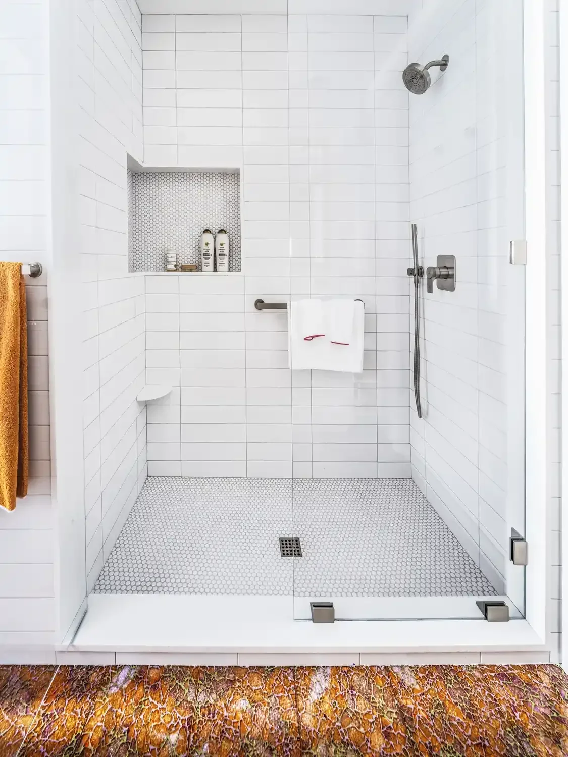Empty walk-in shower with white tiled walls and small hexagonal floor tiles, built-in niche with toiletries, and a yellow towel hanging outside the shower door.