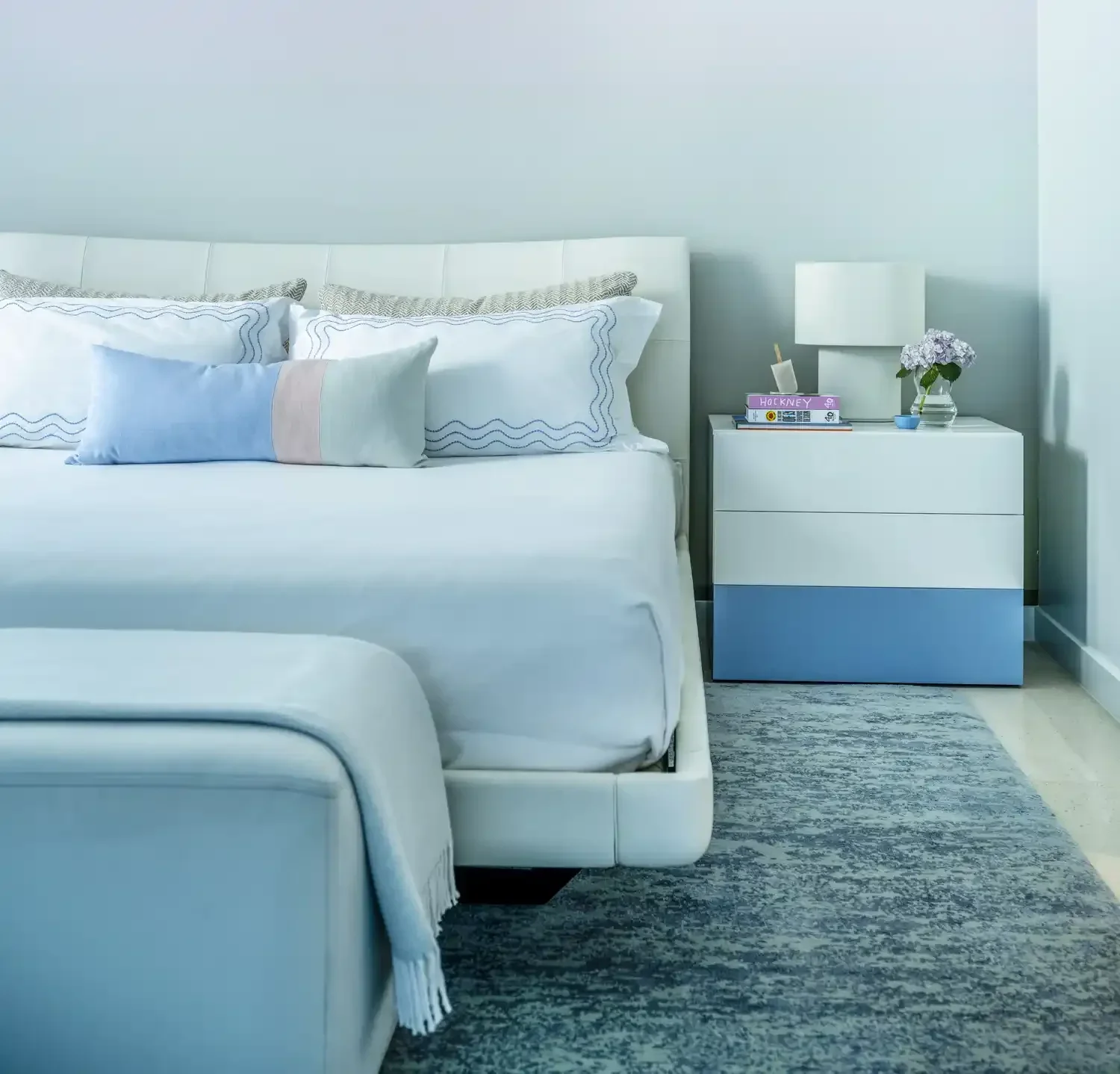 A bright bedroom with a white bed, blue and white pillows, a white nightstand with a white lamp, books, and a small vase with flowers. The floor features a gray patterned rug.