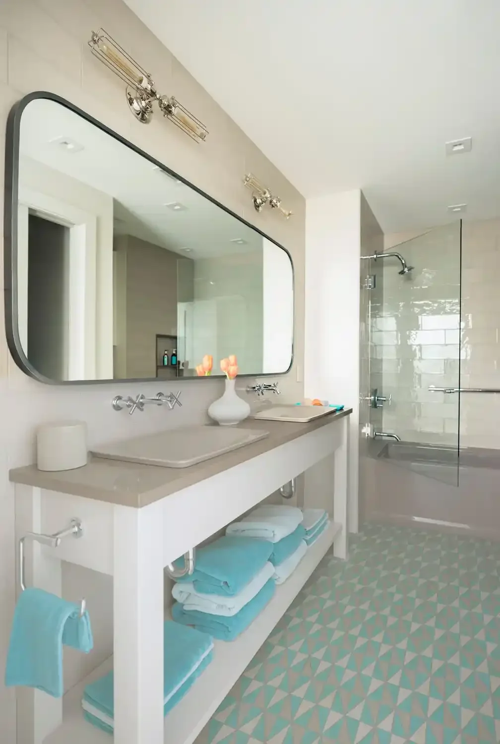 Modern bathroom with a large mirror, white countertop, and shelves with neatly folded blue and white towels. There is a white vase with pink flowers on the counter and a glass-enclosed shower with tiled walls and a showerhead.