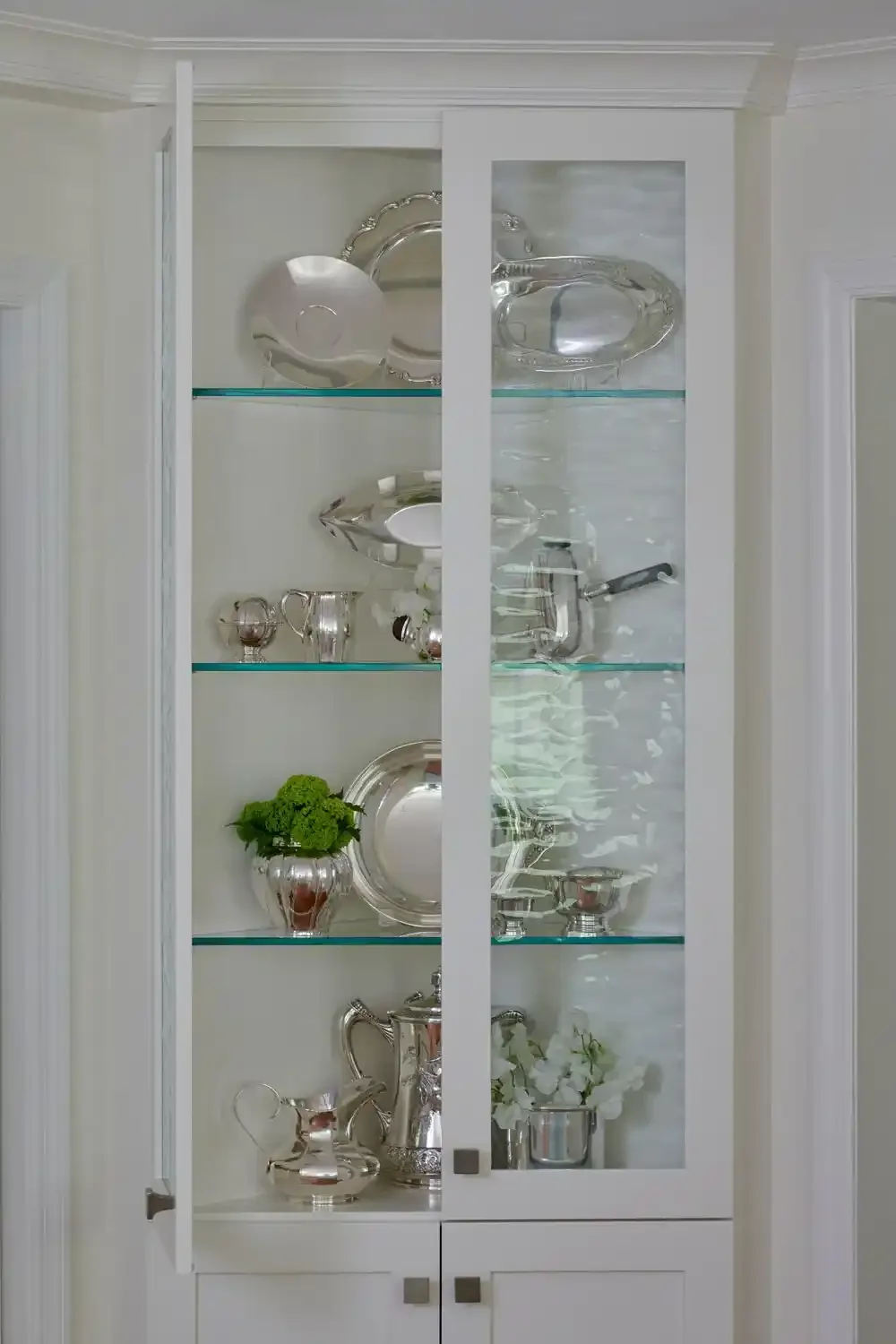 A white display cabinet with glass doors, filled with silver and glass decorative items, pitchers, vases, and flowers.